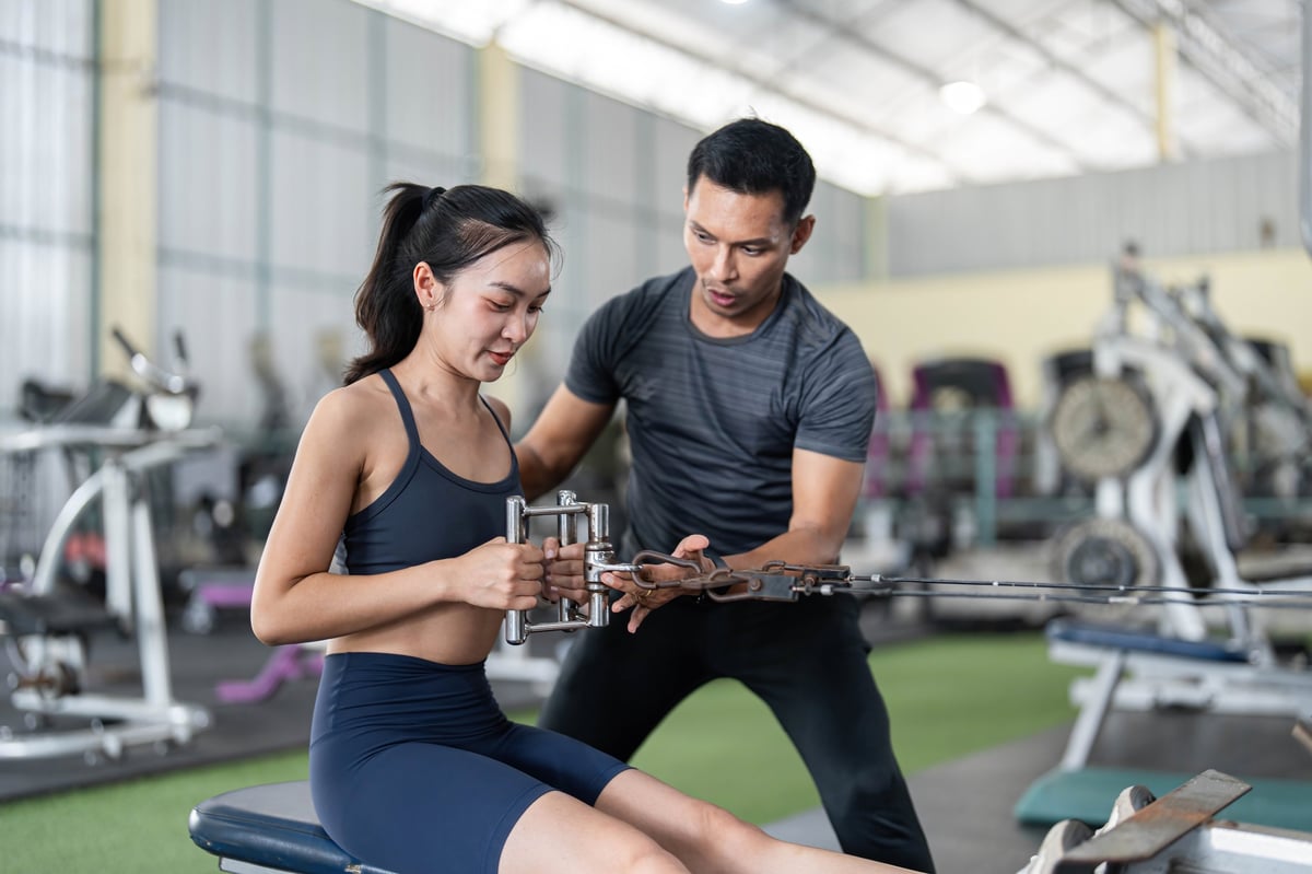A fitness trainer supporting a woman through a strength exercise in a bright gym